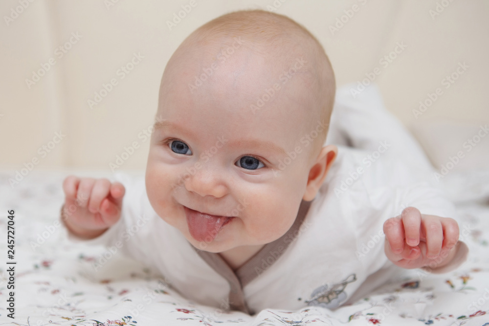 Beautiful five-month blue-eyed baby girl lies on her stomach. Shows tongue, various emotions and a smile on the face. Infant look. Maternal care. Childcare. Close-up. Beautiful five-month blue-eyed baby girl lies on her stomach. Shows tongue, various emotions and a smile on the face. Infant look. Maternal care. Childcare. Close-up.