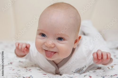 Beautiful five-month blue-eyed baby girl lies on her stomach. Shows tongue, various emotions and a smile on the face. Infant look. Maternal care. Childcare. Close-up.