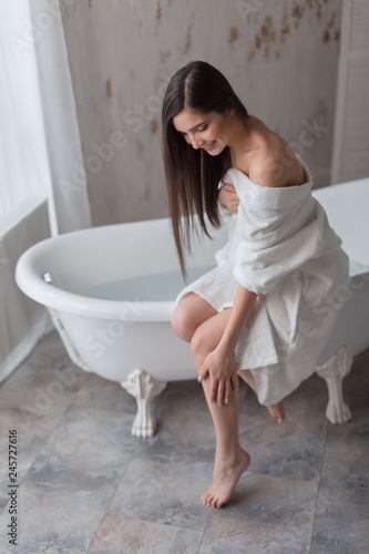 Young woman partially dressed in bathrobe, sits on the edge of bathtub and moisturising with cream her slim long barefoot legs after having bath