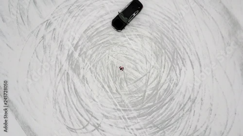 Top view rotation shot sliding and drifting black sport car moving on circle person standing in center. High angle winter race competition surrounded by white snow and ice aerial shot