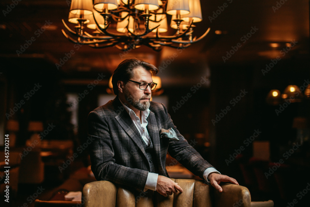 Φωτογραφία Portrait of wealthy prosperious handsome businessman, dressed in tailored three-piece suit sitting in leather arm-chai at elite gentlemen club with lighted multi-lamp chandelier behind