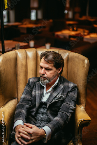 Portrait of fashionable trendy elegant wealthy professional trendsetter mature designer, dressed in tailored plaid jacket, sitting in luxury armchair in living room at home.