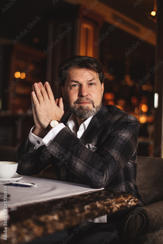 Mature confident businessman in formal bespoke suit looking at camera with serious pensive expression sitting in dark restaurant with artificial lighting and luxurious interior. Mature confident businessman in formal bespoke suit looking at camera with serious pensive expression sitting in dark restaurant with artificial lighting and luxurious interior.