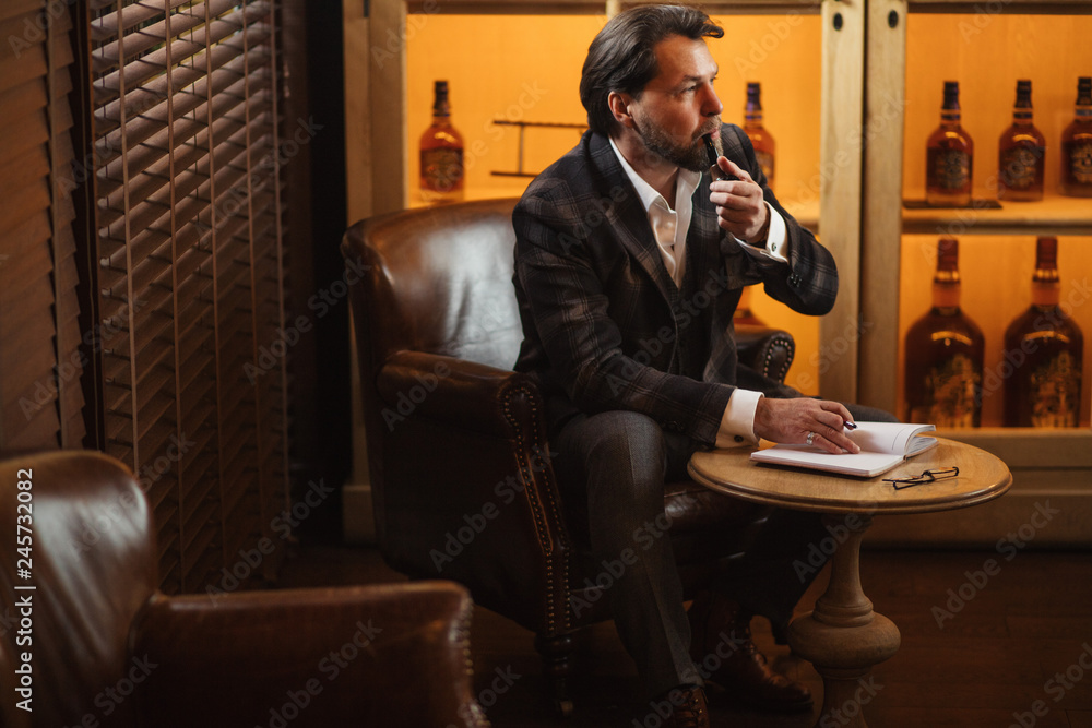 Portrait of busy and hard-working fashion designer smoking pipe and making notes into his diary at his laxurious office.