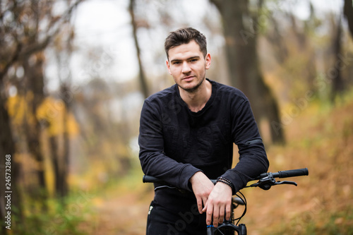 Fototapeta Naklejka Na Ścianę i Meble -  Young pretty athletic man standing with bicycle in colorful autumn park. Fall season background. Male cyclist on the road with fallen leaves
