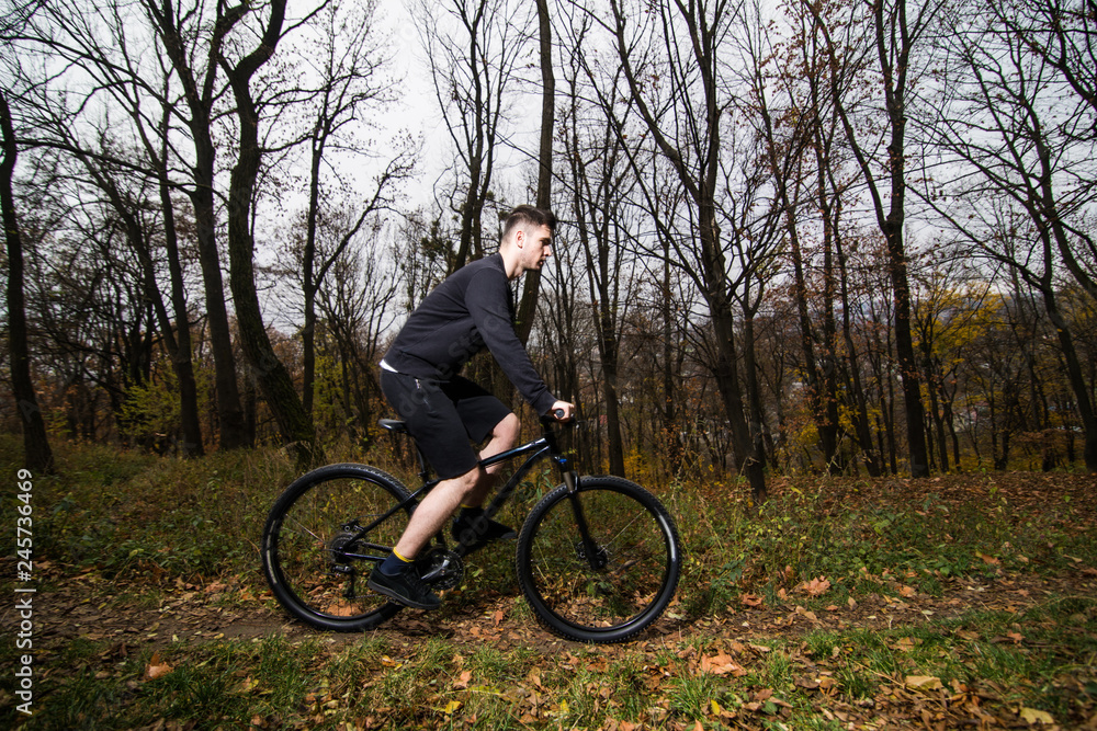Handsome male biker in professional sportswear while riding bike down park alley on autumn day. Sportsman training thinking about future win in contest. Handsome male biker in professional sportswear while riding bike down park alley on autumn day. Sportsman training thinking about future win in contest.