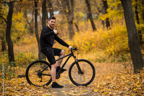 Handsome man biker in professional sportswear looking to side while riding bike down park alley on autumn day. Sportsman training thinking about future win in contest