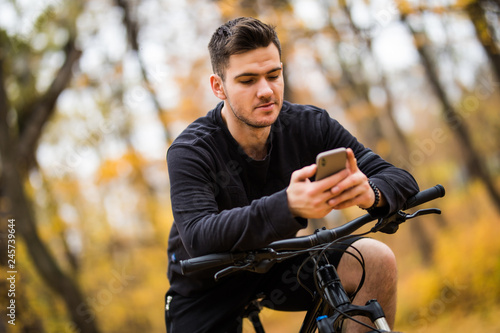 Young sportsman riding bicycle holding phone, sunny autumn park