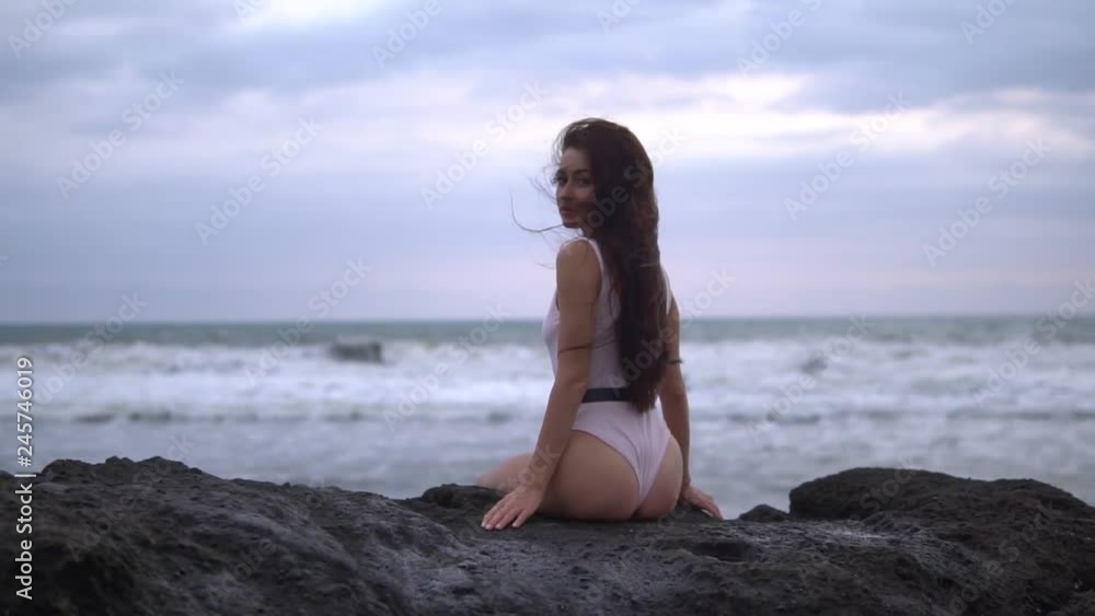 young pretty female with long dark curly hair in white swimwear sits on rock near ocean young pretty female with long dark curly hair in white swimwear sits on rock near ocean