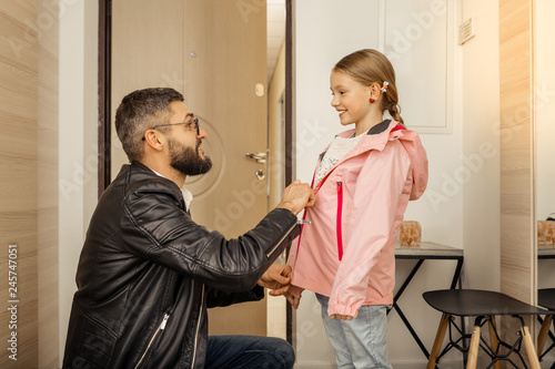Tablou pe pânză Handsome dark-haired bearded man in eyewear zipping a jacket on his daughter