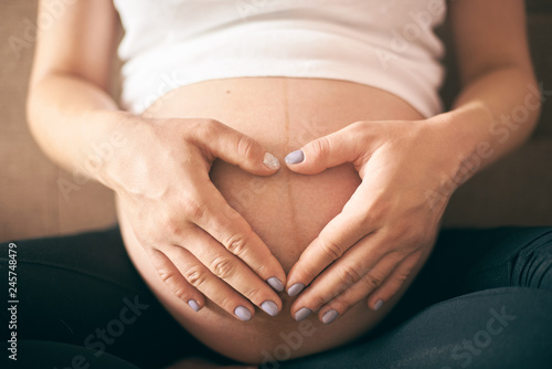Pregnant woman in white shirt sitting at home and embracing big belly. Selective focus of female hands showing heart near tummy. Future mother expecting baby.