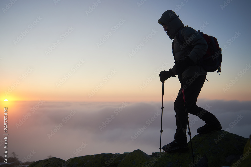 Tourist hiker in warm clothing with backpack and trekking poles climbing on rocky mountain peak on foggy cloudy landscape, misty blue sky and raising bright orange sun at dawn copy space background.