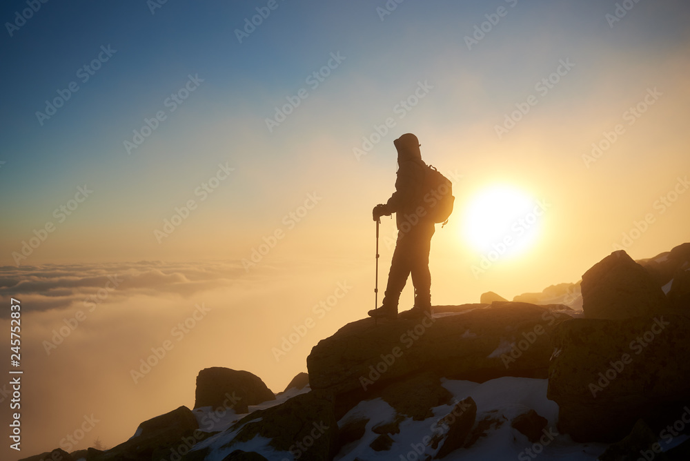 Silhouette of tourist hiker with backpack and hiking sticks resting on the top of rocky snowy mountain on copy space background of bright white raising sun and misty orange blue sky at dawn.