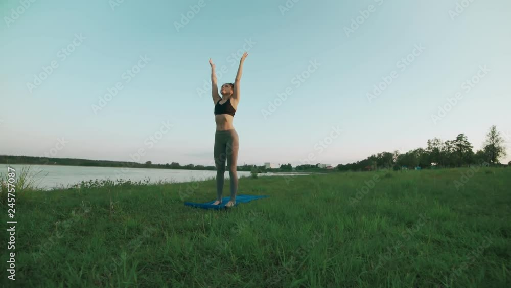 Woman doing yoga in quiet scenery. Beautiful young girl wearing sports clothes doing yoga in morning at lake, sun salutation on blue yoga mat. Slide left with copyspace Woman doing yoga in quiet scenery. Beautiful young girl wearing sports clothes doing yoga in morning at lake, sun salutation on blue yoga mat. Slide left with copyspace