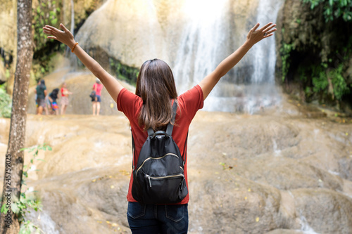 Rear view of Asian woman standing in front of waterfall with her hands raised. 