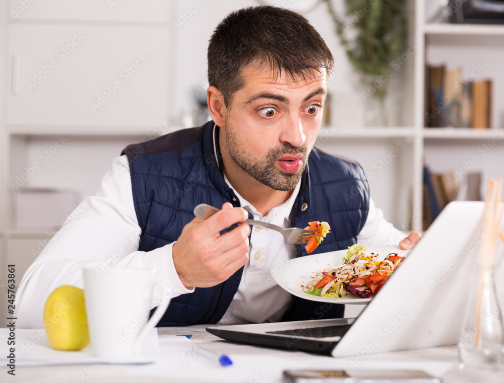 Portrait of man eating vegetable salad at table with laptop