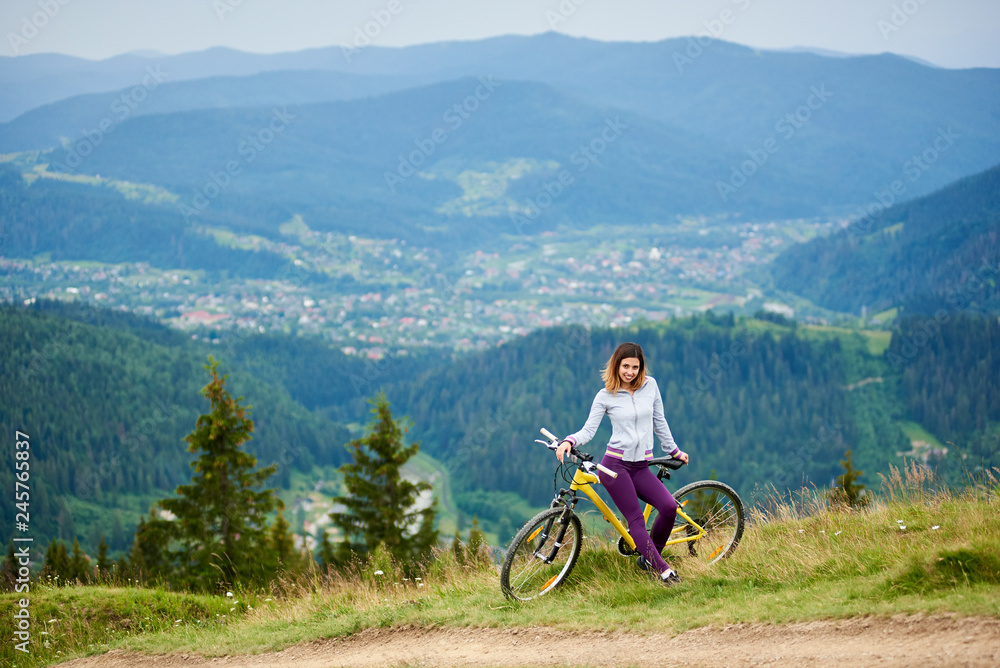 Happy female cyclist resting on yellow bicycle in the mountains on cloudy evening. Mountains, forests and small city on the blurred background. Outdoor sport activity, lifestyle concept