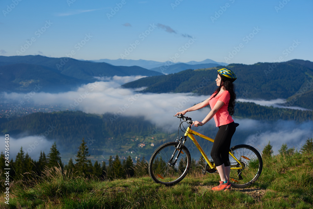 Sporty female biker with yellow bicycle in the mountains, wearing helmet and red red t-shirt in the morning. Foggy mountains, forests on the blurred background. Outdoor sport activity. Copy space
