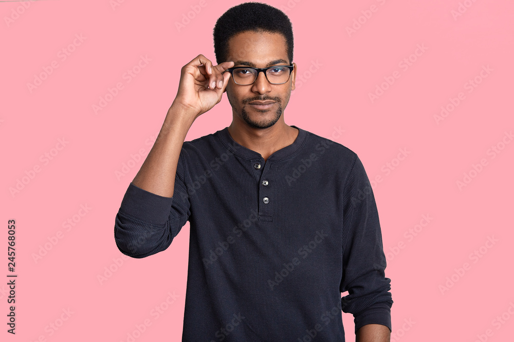Horizontal shot of serious black man keeps hand on frame of spectacles, has self confident expression, wears black jumper, has dark healthy skin, isolated over pink background, listens something Horizontal shot of serious black man keeps hand on frame of spectacles, has self confident expression, wears black jumper, has dark healthy skin, isolated over pink background, listens something