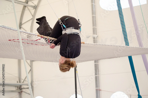 An adult female lands on a net, preparing to dismount at a on a flying trapeze school at an indoor gym. The woman is an amateur trapeze artist.