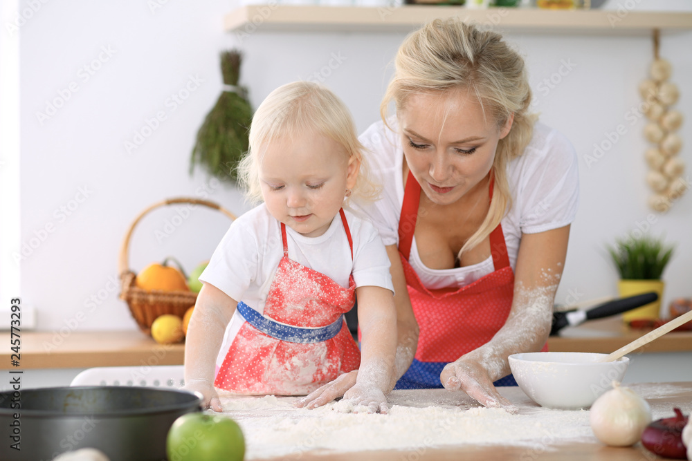 Little girl and her blonde mom in red aprons playing and laughing while kneading the dough in kitchen. Homemade pastry for bread, pizza or bake cookies. Family fun and cooking concept Little girl and her blonde mom in red aprons playing and laughing while kneading the dough in kitchen. Homemade pastry for bread, pizza or bake cookies. Family fun and cooking concept