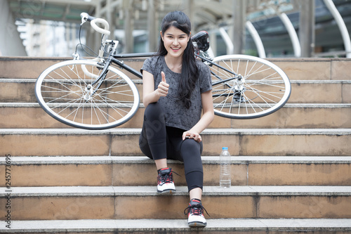 asian young sport woman in sportswear sitting on the stairs with bicycle and showing thumbs up . girl taking a rest and relaxing on street in urban city . workout exercising