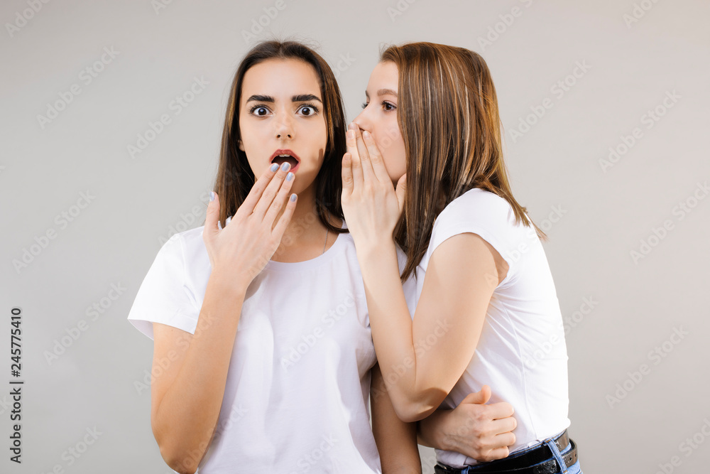 Two beautiful female dressed in white shirts gossiping. Two cute girls posing at the camera while one is whispering a secret on another's ear while another opens hers mouth amazed . Two beautiful female dressed in white shirts gossiping. Two cute girls posing at the camera while one is whispering a secret on another's ear while another opens hers mouth amazed .