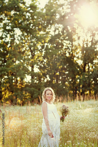 Beautiful and happy pregnant woman in a white dress on the nature in the summer, around the trees and flowers.