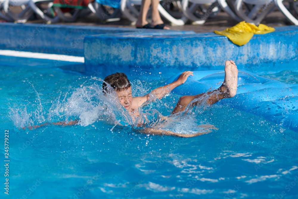 Cute European boy is using blue air mattress, while having fun in hotel’s swimming pool. He is diving into the water.