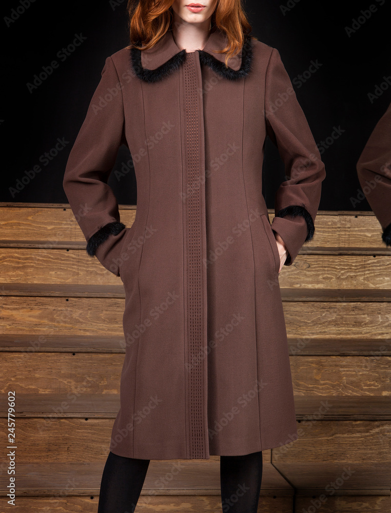 Woman in coat and photomodel posing. Wooden floor and stairs. Black background with mirror and reflection. Fashion and design, luxury. Coffee-colored coat