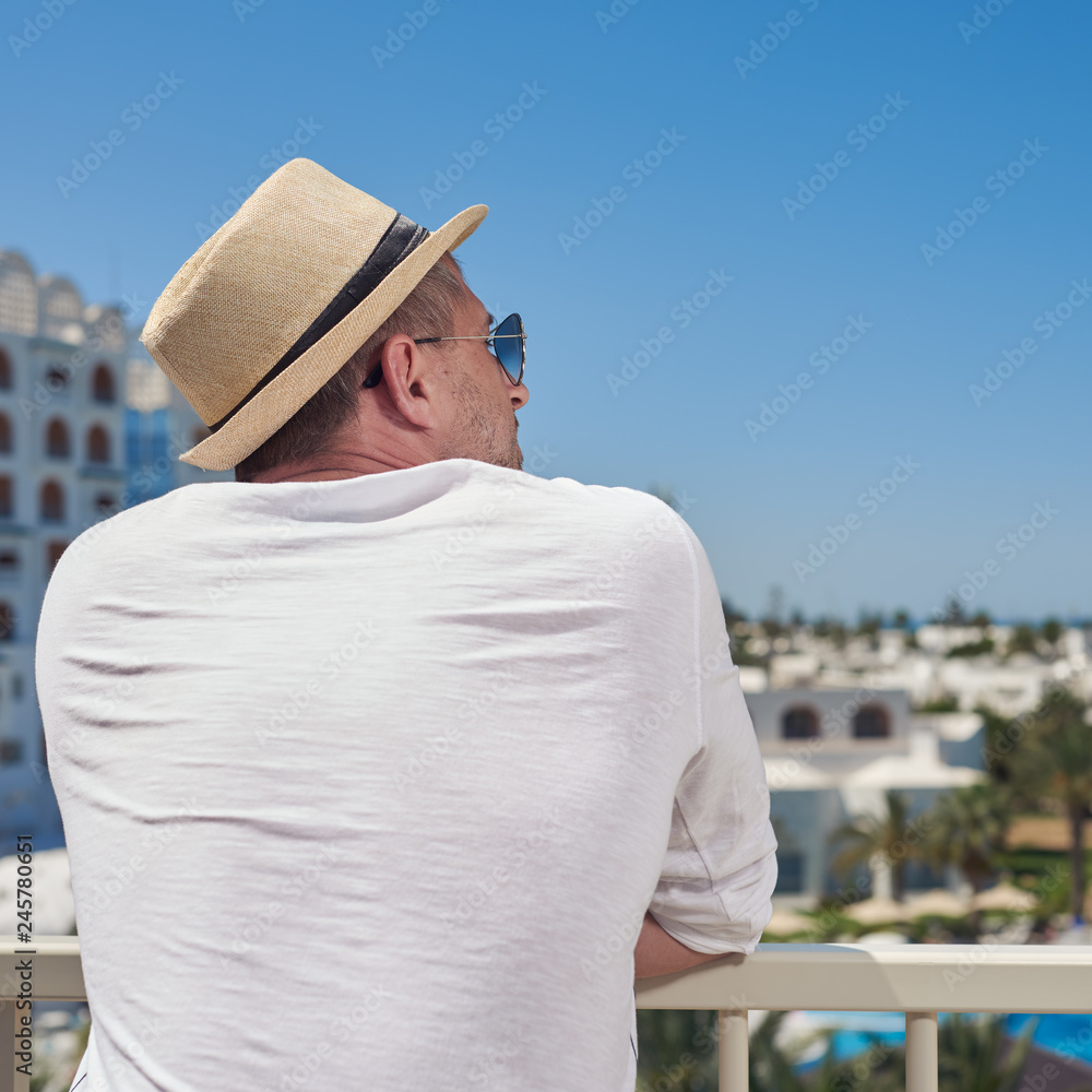 Attractive European man in sunhat is observing wonderful hotel’s view. He is relaxing and enjoying his summer holidays.