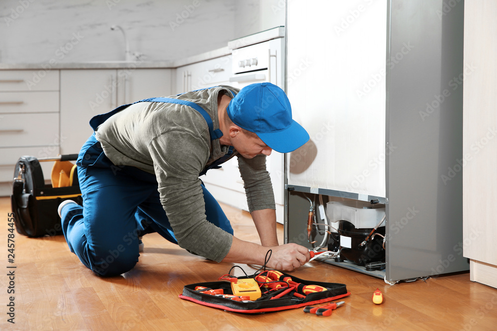 Male technician in uniform repairing refrigerator indoors - obrazy, fototapety, plakaty 