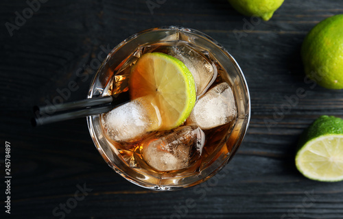 Glass of cocktail with cola, ice and cut lime on wooden background, top view photo