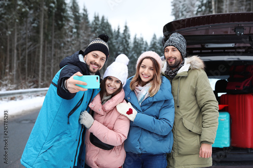 Friends taking selfie near open car trunk on road. Winter vacation