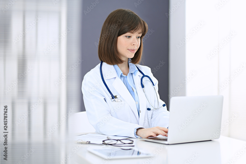 Doctor typing on laptop computer while sitting at the white table in hospital office. Physician at work. Medicine and healthcare concept Doctor typing on laptop computer while sitting at the white table in hospital office. Physician at work. Medicine and healthcare concept