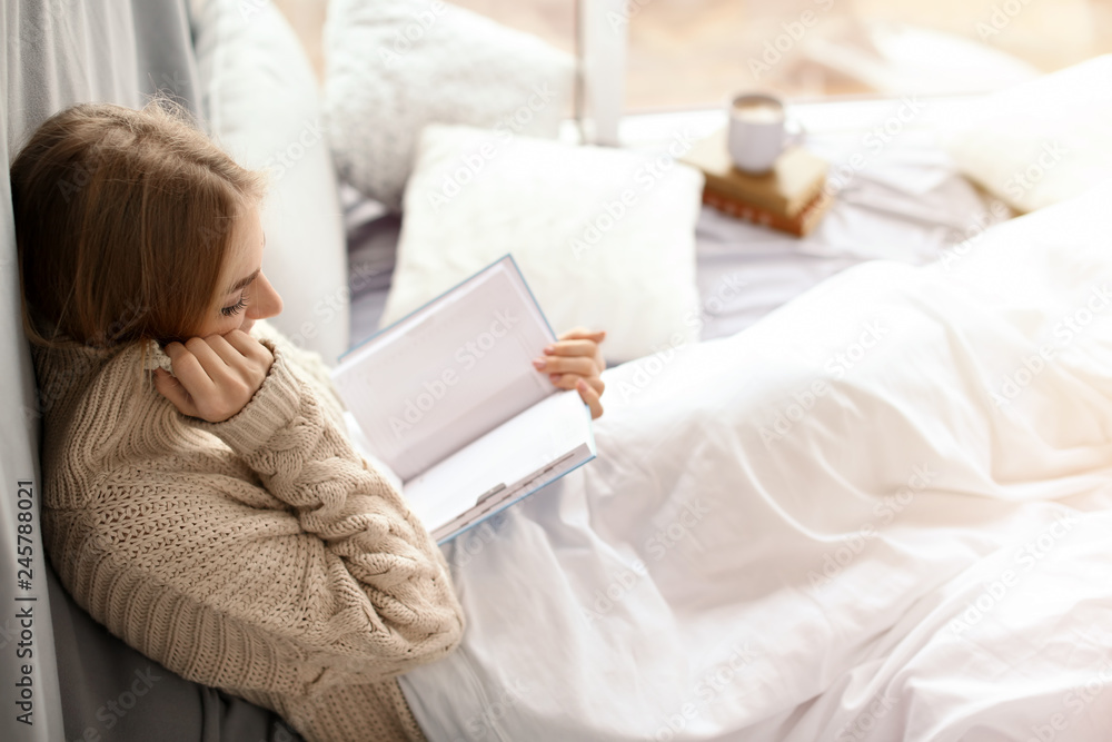 Beautiful young woman in knitted sweater sitting and reading book near window at home. Winter atmosphere Beautiful young woman in knitted sweater sitting and reading book near window at home. Winter atmosphere