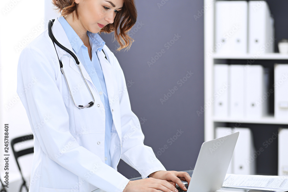 Happy doctor woman at work. Portrait of female physician using laptop computer while standing near reception desk at clinic or emergency hospital. Medicine concept Happy doctor woman at work. Portrait of female physician using laptop computer while standing near reception desk at clinic or emergency hospital. Medicine concept