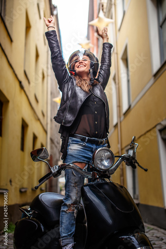 Cheerful hipster girl wearing a leather jacket and ripped jeans standing in a black classic scooter on the old narrow street © Fxquadro