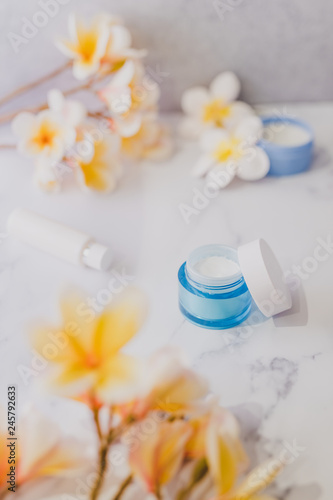 group of skincare products including moisturiser scrub and hand cream pots on marble table with exotic frangipani flowers