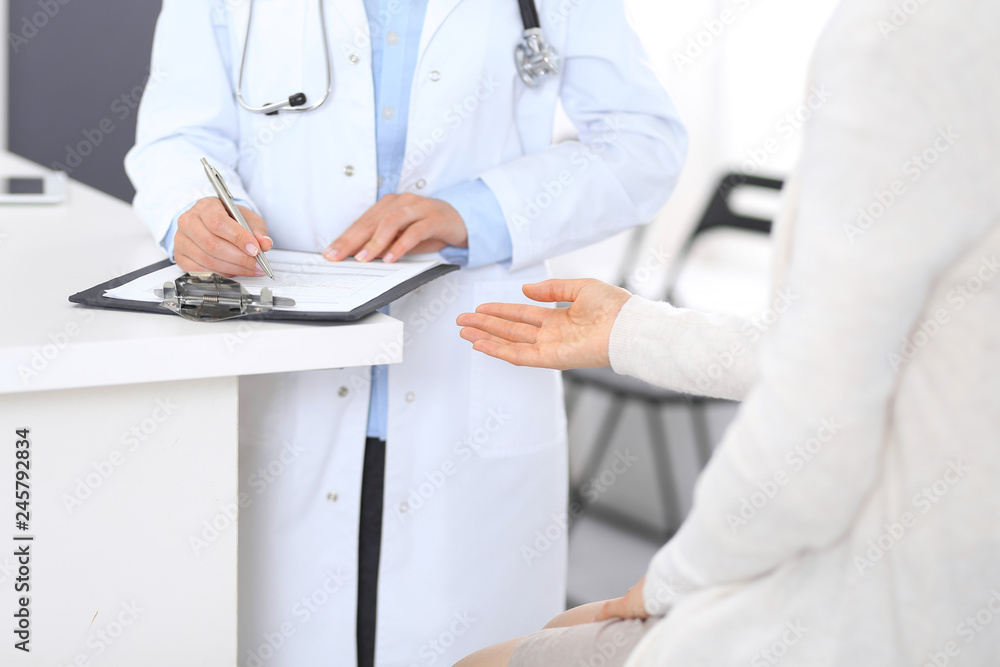 Unknown doctor and female patient discussing something while standing near reception desk in emergency hospital. Physician at work in clinic. Medicine and health care concept Unknown doctor and female patient discussing something while standing near reception desk in emergency hospital. Physician at work in clinic. Medicine and health care concept