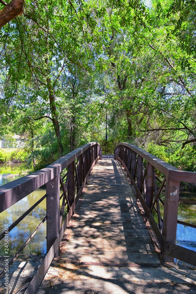 Views of Jordan River Trail Pedestrian and Train Track Bridge with surrounding trees, Russian Olive, cottonwood and muddy stream along the Wasatch Front Rocky Mountains, in Salt Lake City, Utah.