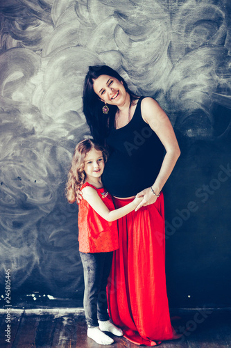 Portrait of pregnant mother and happy cheerful curly daughetr hugging on black background in red and black colors. Black background. Pregnancy , family, happiness concept photo