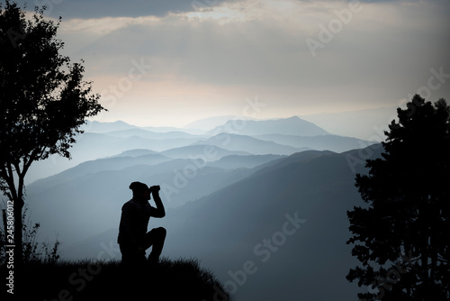 silhouette of a man on the top of a mountain while looking at the landscape with binoculars