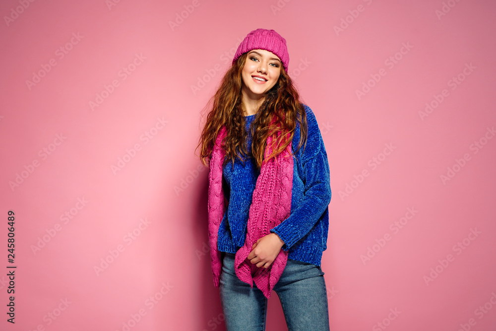 Modern young woman wearing blue sweater and pink hat, and scarf posing, making funny facial expression. Retouched, studio portrait. Modern young woman wearing blue sweater and pink hat, and scarf posing, making funny facial expression. Retouched, studio portrait.