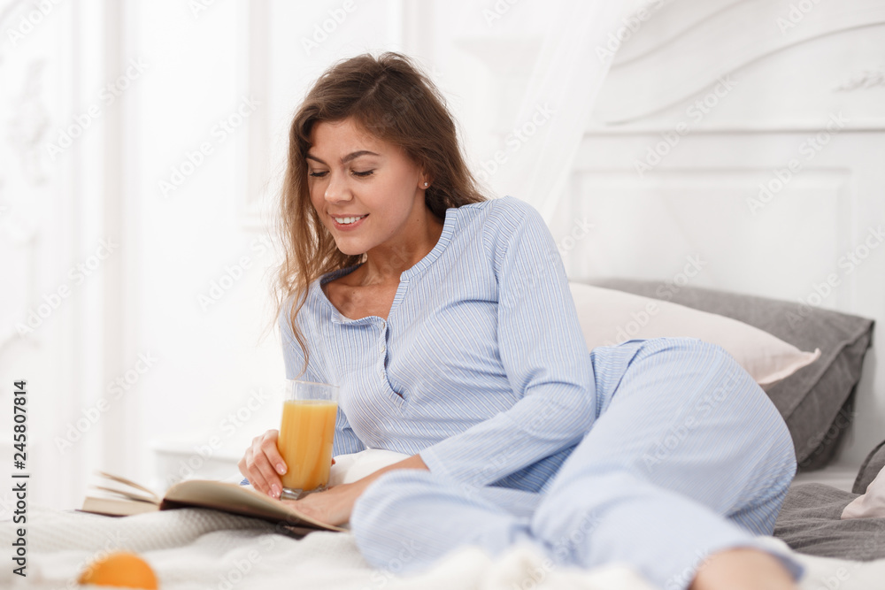 Pretty brunette girl dressed in the light-blue pajama lays on the bed with glass of fresh juice and reads book in the cozy light room Pretty brunette girl dressed in the light-blue pajama lays on the bed with glass of fresh juice and reads book in the cozy light room