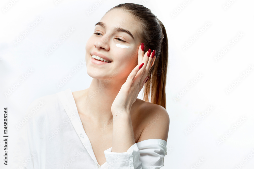 Smiling young brown-haired girl without makeup dressed in white clothes puts a cream on her face on the white background Smiling young brown-haired girl without makeup dressed in white clothes puts a cream on her face on the white background