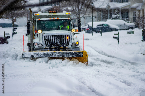 White truck with yellow snow plow blade clearing streets after heavy snowfall in urban area with driver wearing yellow safety vest