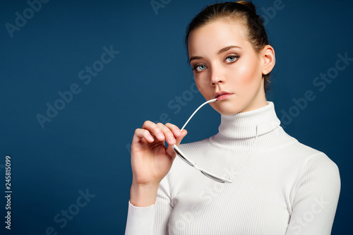 Portrait of a beautiful young girl in white knitted golf and sunglasses on a blue background