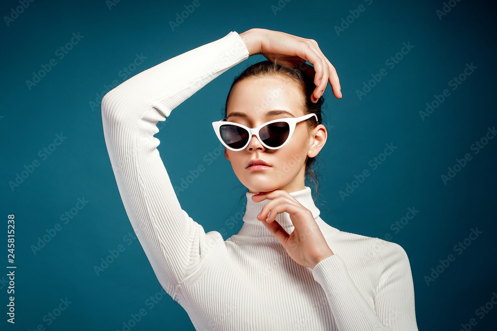 Portrait of a beautiful young girl in white knitted golf and sunglasses on a blue background Portrait of a beautiful young girl in white knitted golf and sunglasses on a blue background