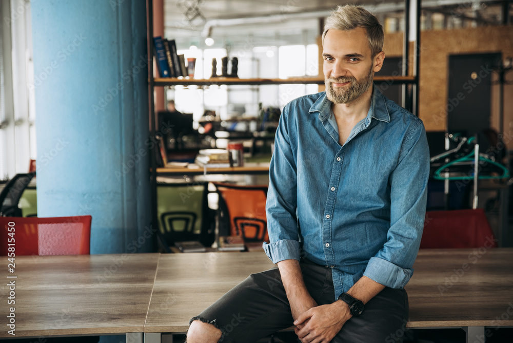 Portrait of a stylish modern guy in a blue shirt and with beard who sits in the office on the table Portrait of a stylish modern guy in a blue shirt and with beard who sits in the office on the table